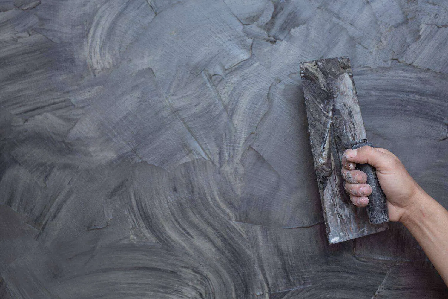 Hand holding trowel applying dark textured plaster to wall surface.