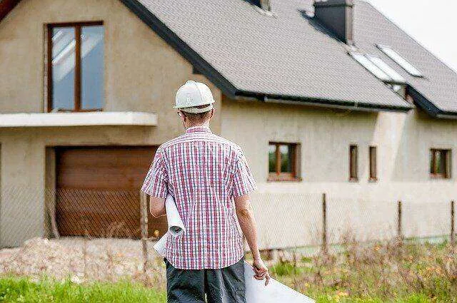 Person in hard hat holding blueprints in front of a modern stucco house.