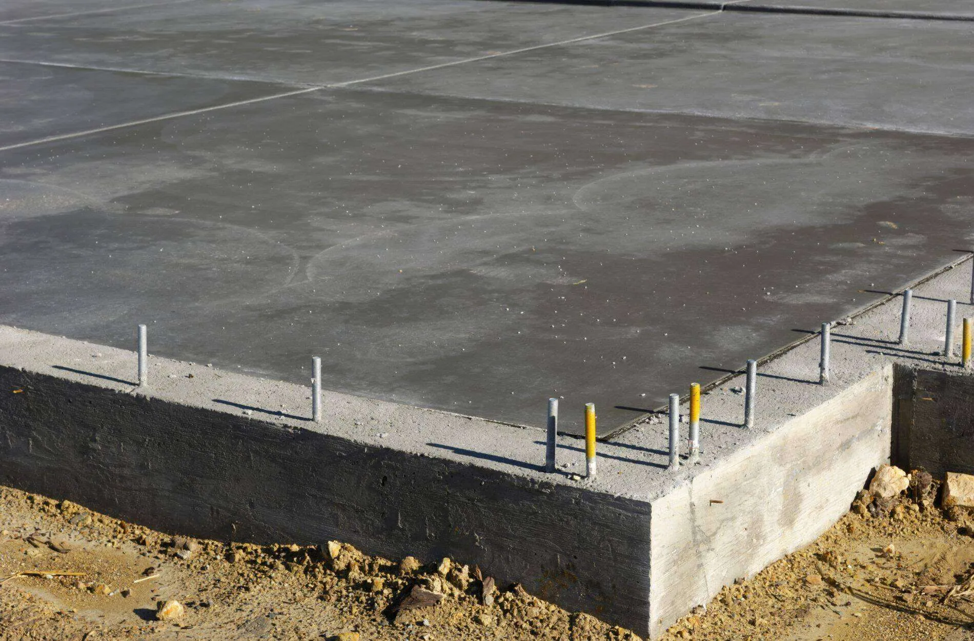 Concrete foundation slab with metal rods on a sandy construction site.