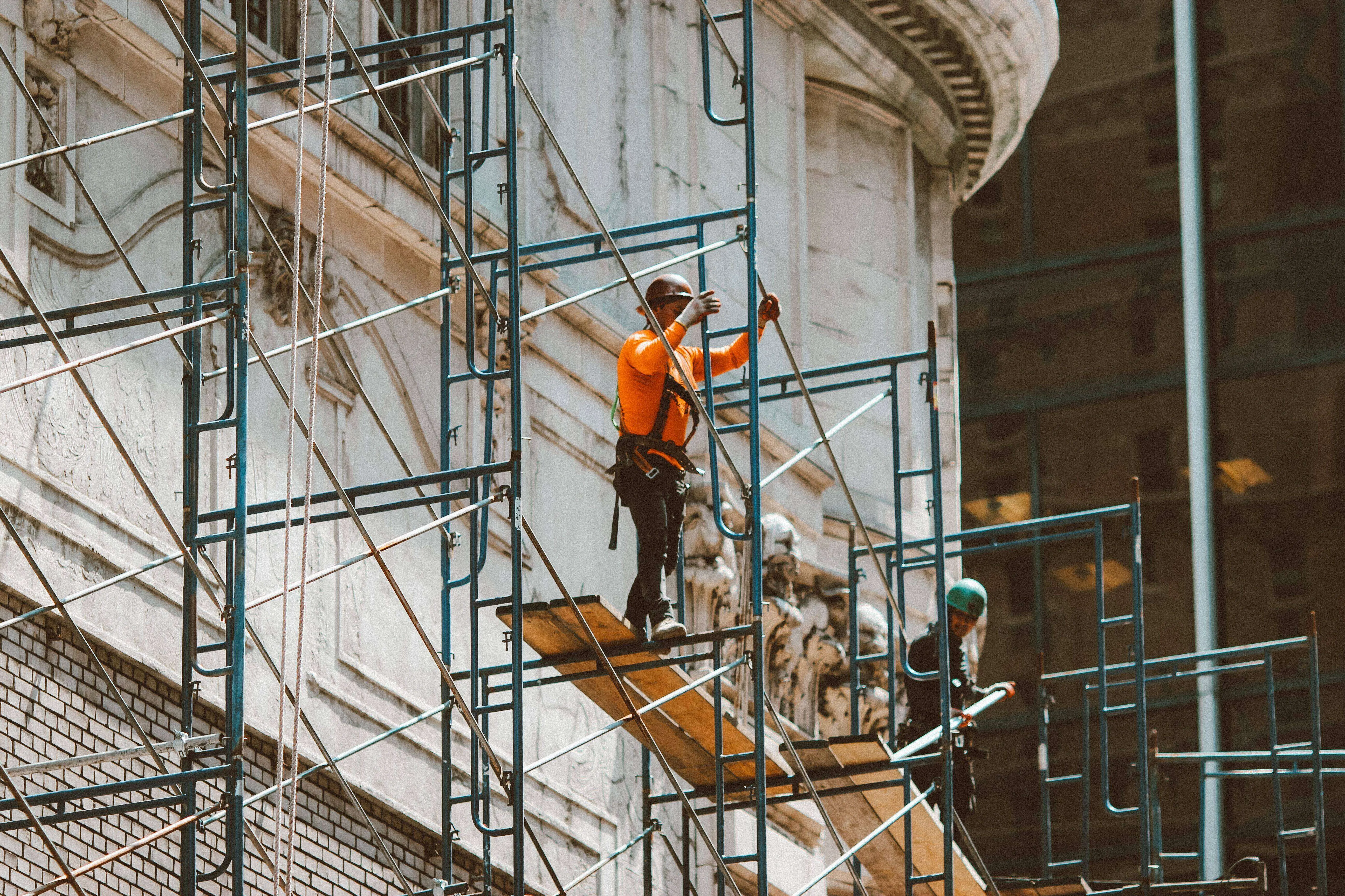 Two workers on scaffolding in front of a historic building facade, wearing safety gear.
