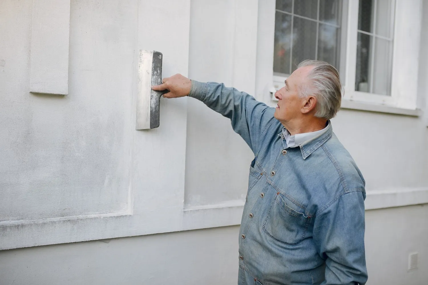 Man in denim shirt applying plaster to an exterior wall with a trowel.