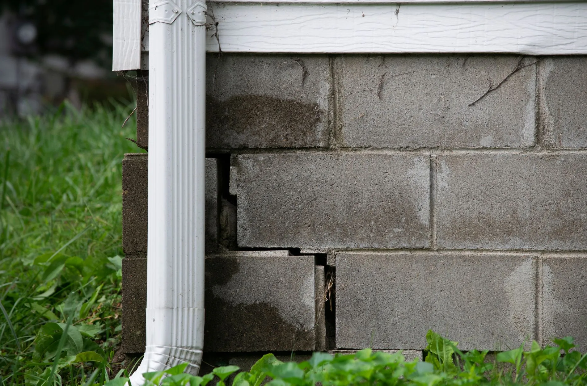 Foundation wall with cracks and water stains, downspout attached, grass in foreground.