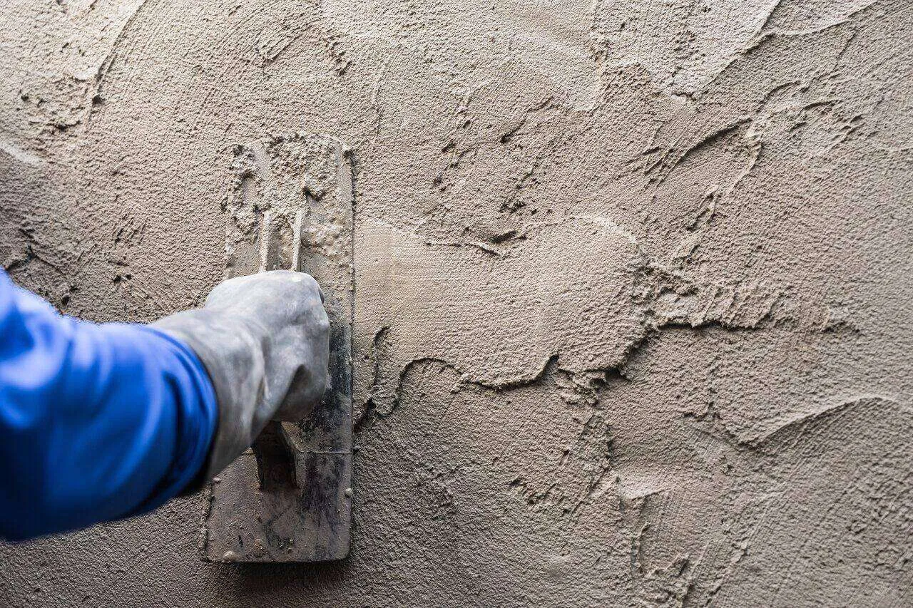 Close-up of a hand plasterer using a trowel to apply cement on a wall, showing rough texture.