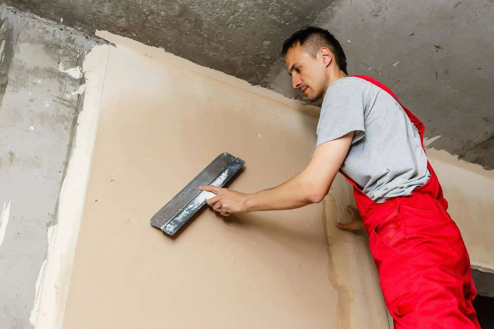 Worker in red overalls applying plaster to a wall with a trowel in an interior setting.