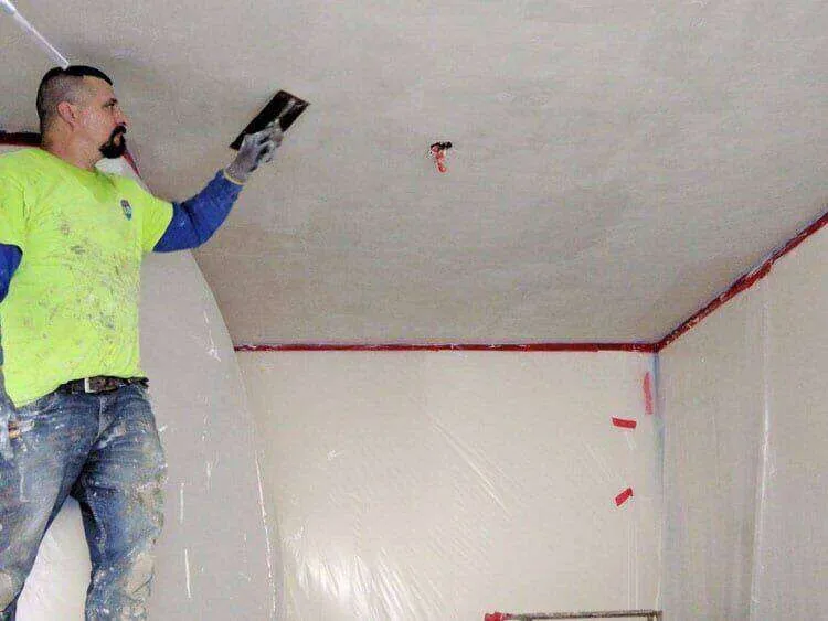 Worker applying plaster to a ceiling in a room covered with protective plastic sheeting.