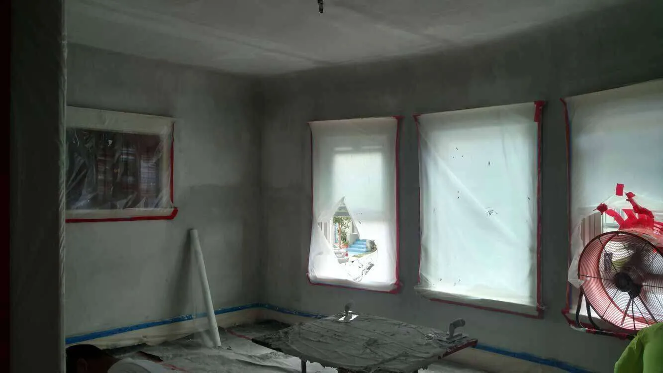 Room with plastered walls, windows covered in plastic, and a fan during renovation.