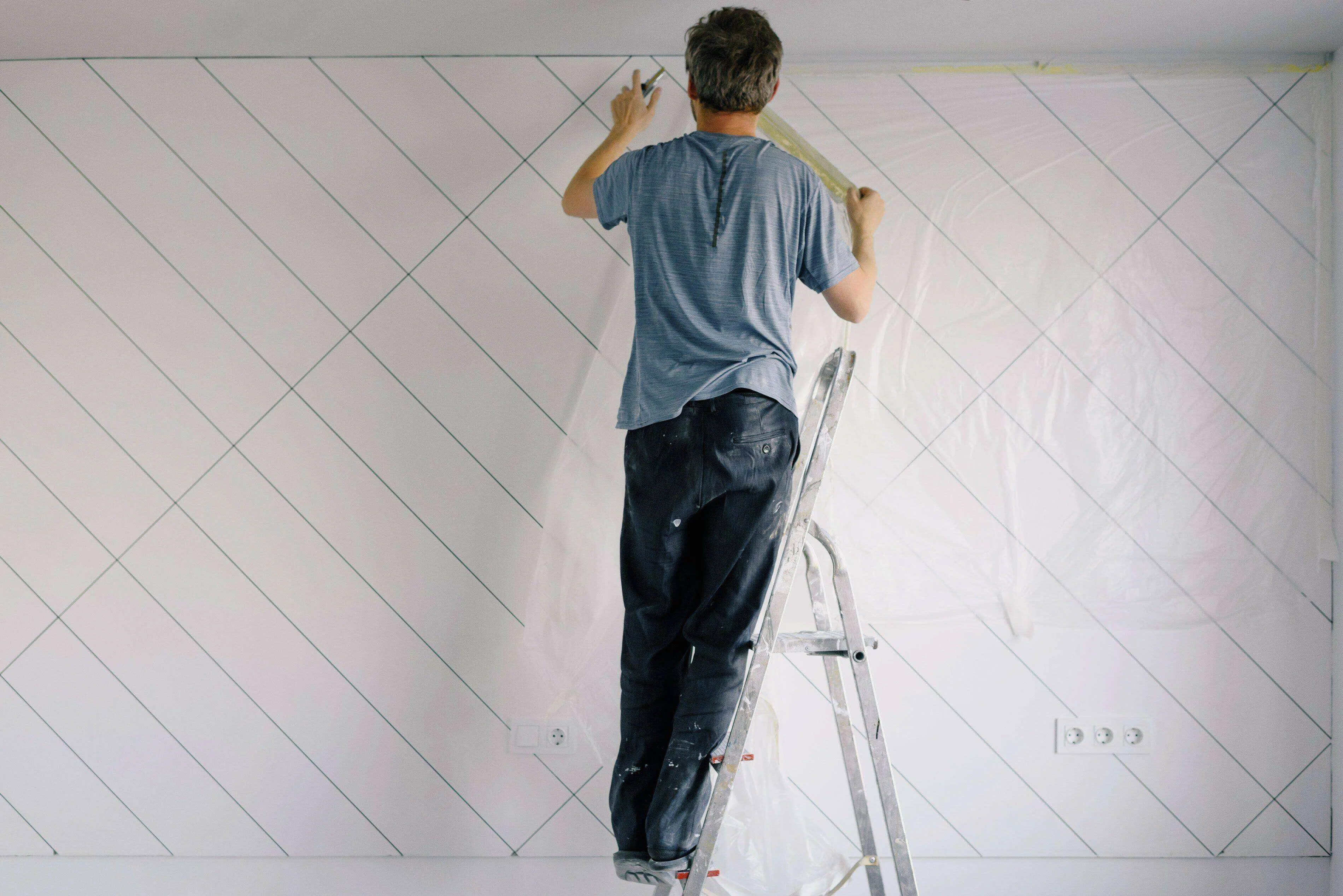 Person on ladder applying tape to a wall with diagonal lines and plastic sheeting.