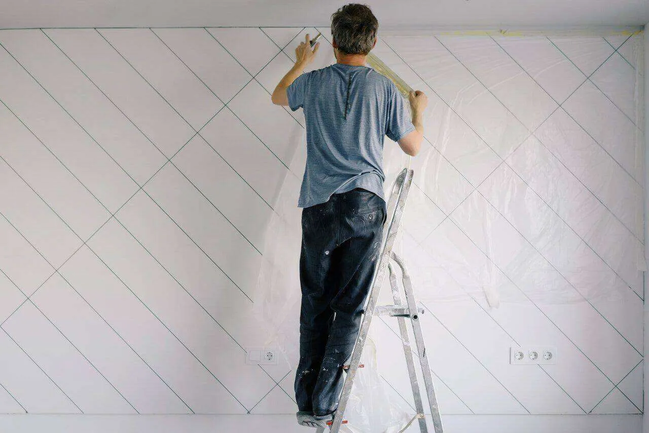 Person on ladder applying masking tape to a wall with grid pattern and plastic sheeting.
