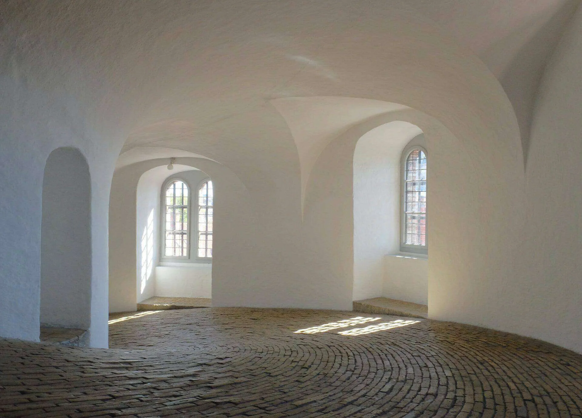 Interior space with white plastered walls, arched windows, and a curved brick floor.