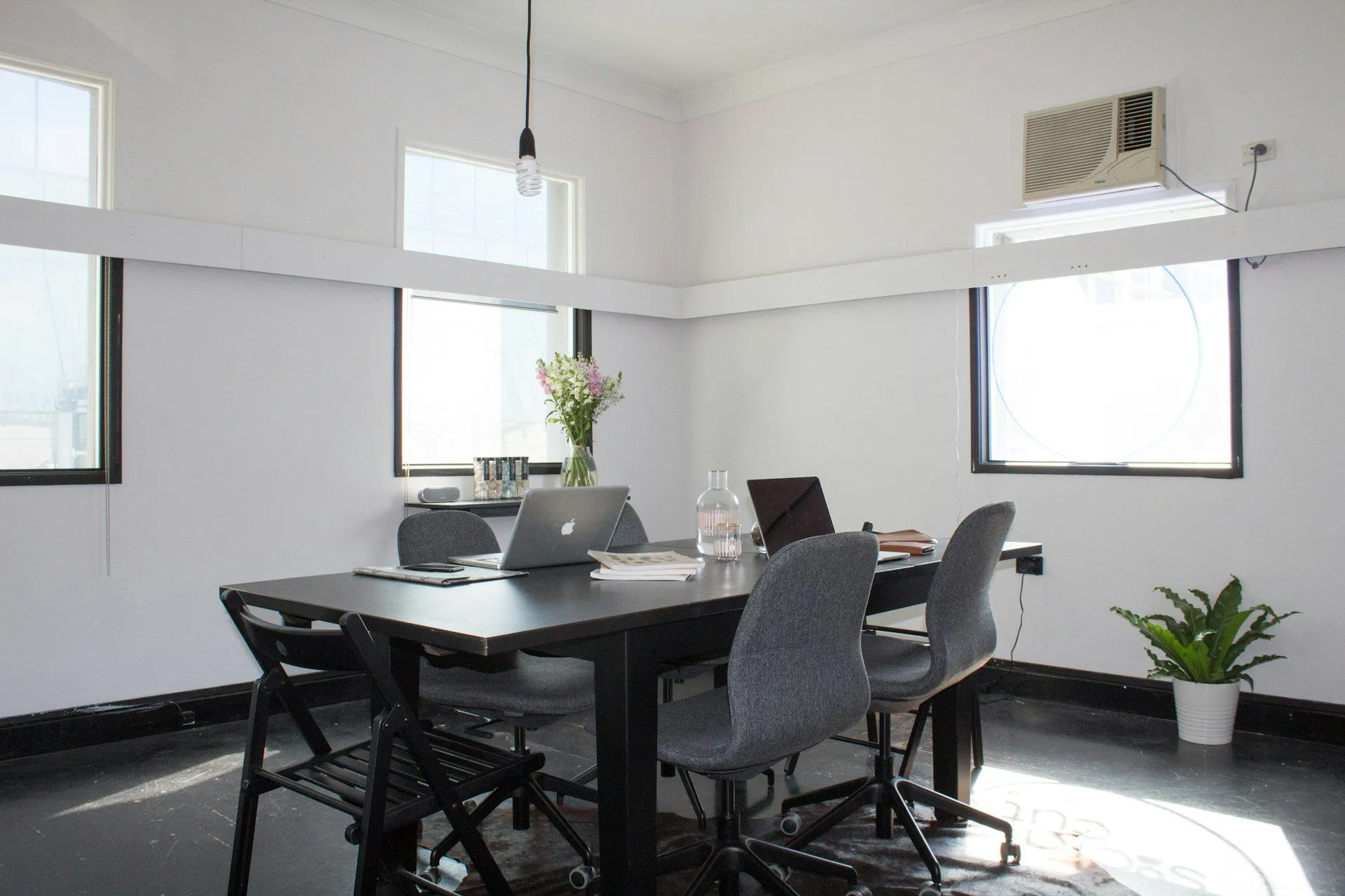 Office interior with table, chairs, laptops, and natural light from large windows.