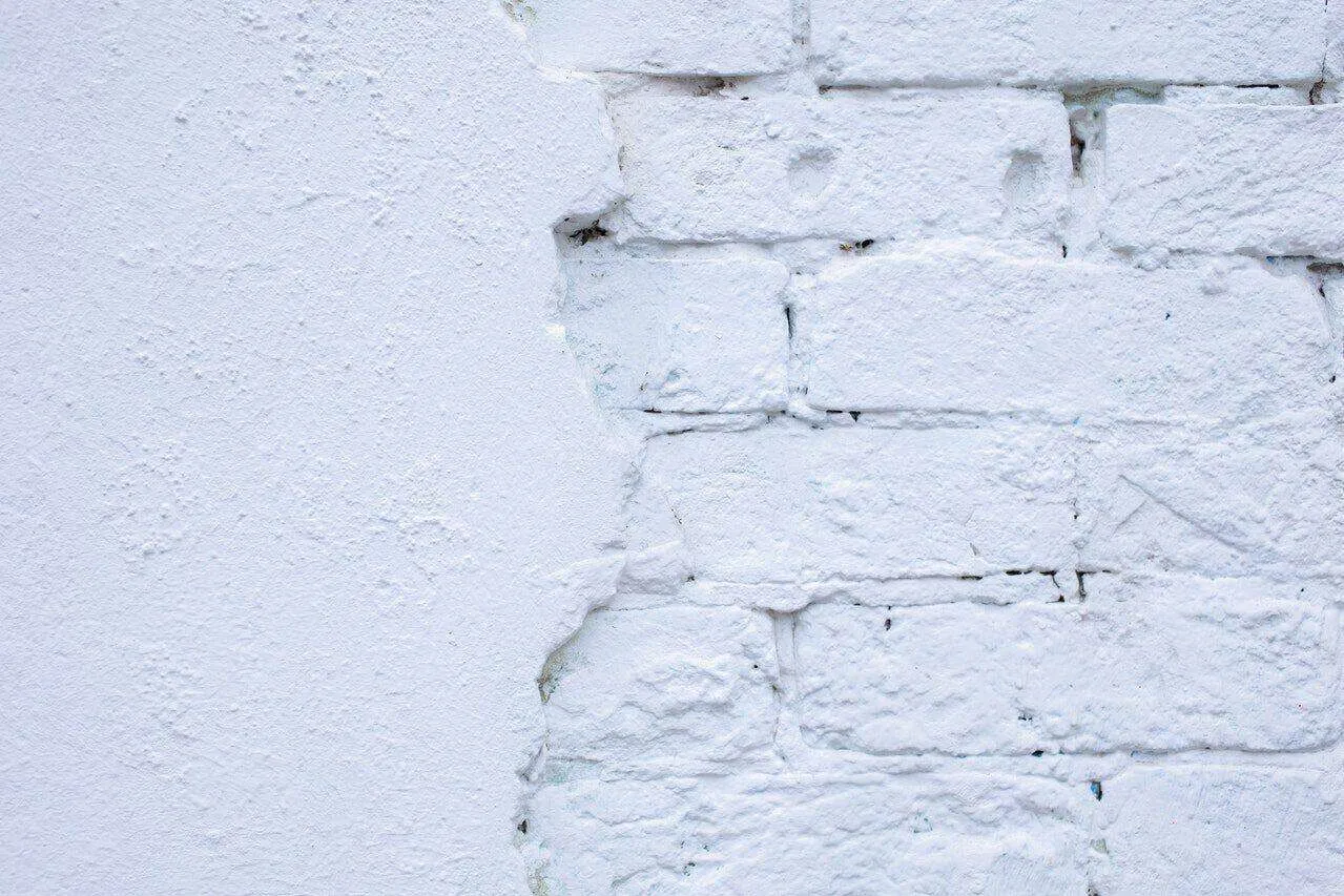 Close-up of white plaster peeling off a white brick wall, revealing bricks underneath.