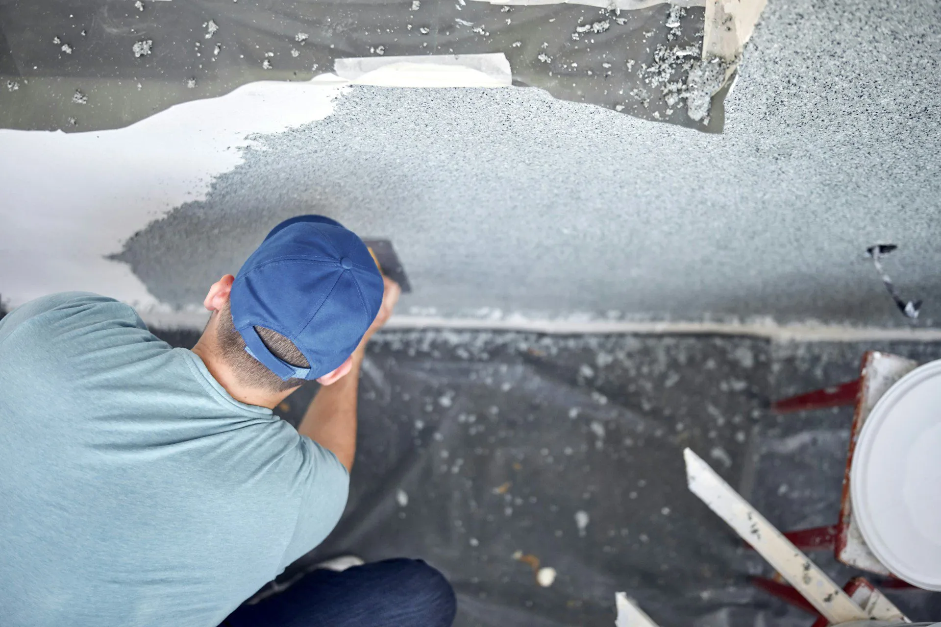 Person in blue cap applying plaster to a wall with protective sheets and tools around.
