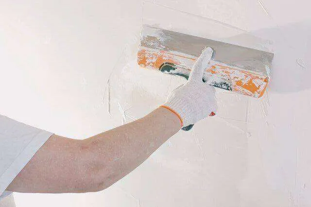 Person applying plaster to a wall with a trowel, wearing a glove.