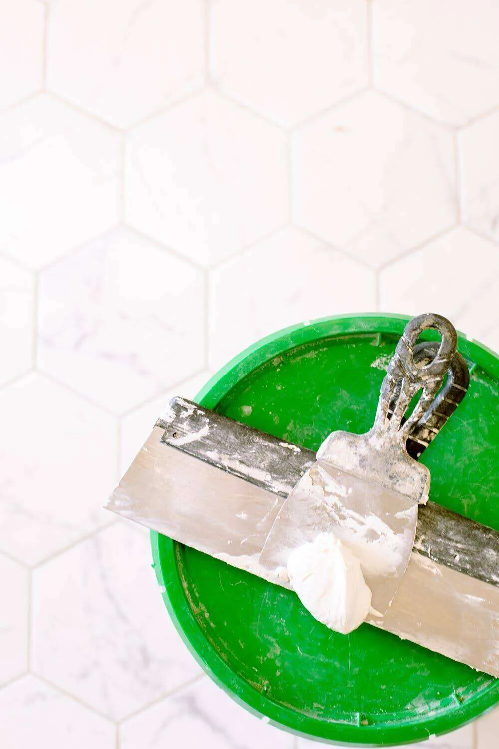 Green bucket with plastering trowel on top, against white hexagonal tile background.