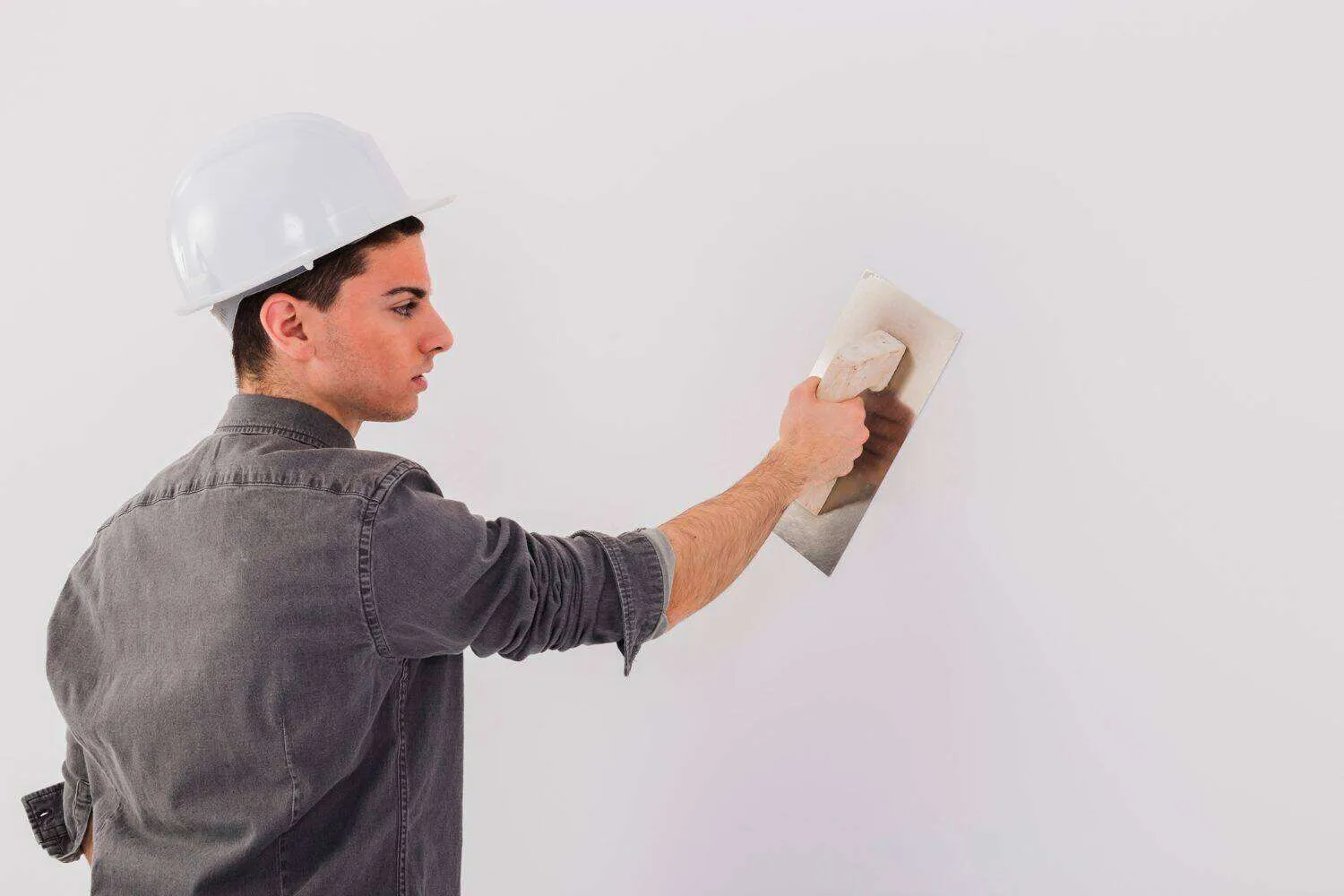 Person in hard hat applying plaster to a smooth white wall with a trowel.
