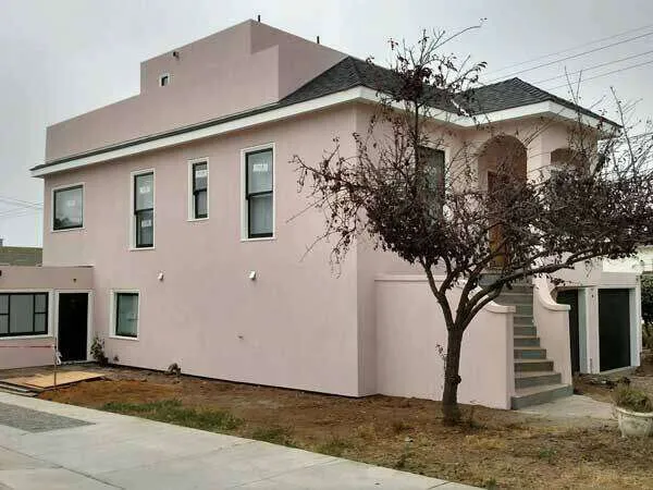 Two-story pink stucco house with black-framed windows and a bare yard with a tree.