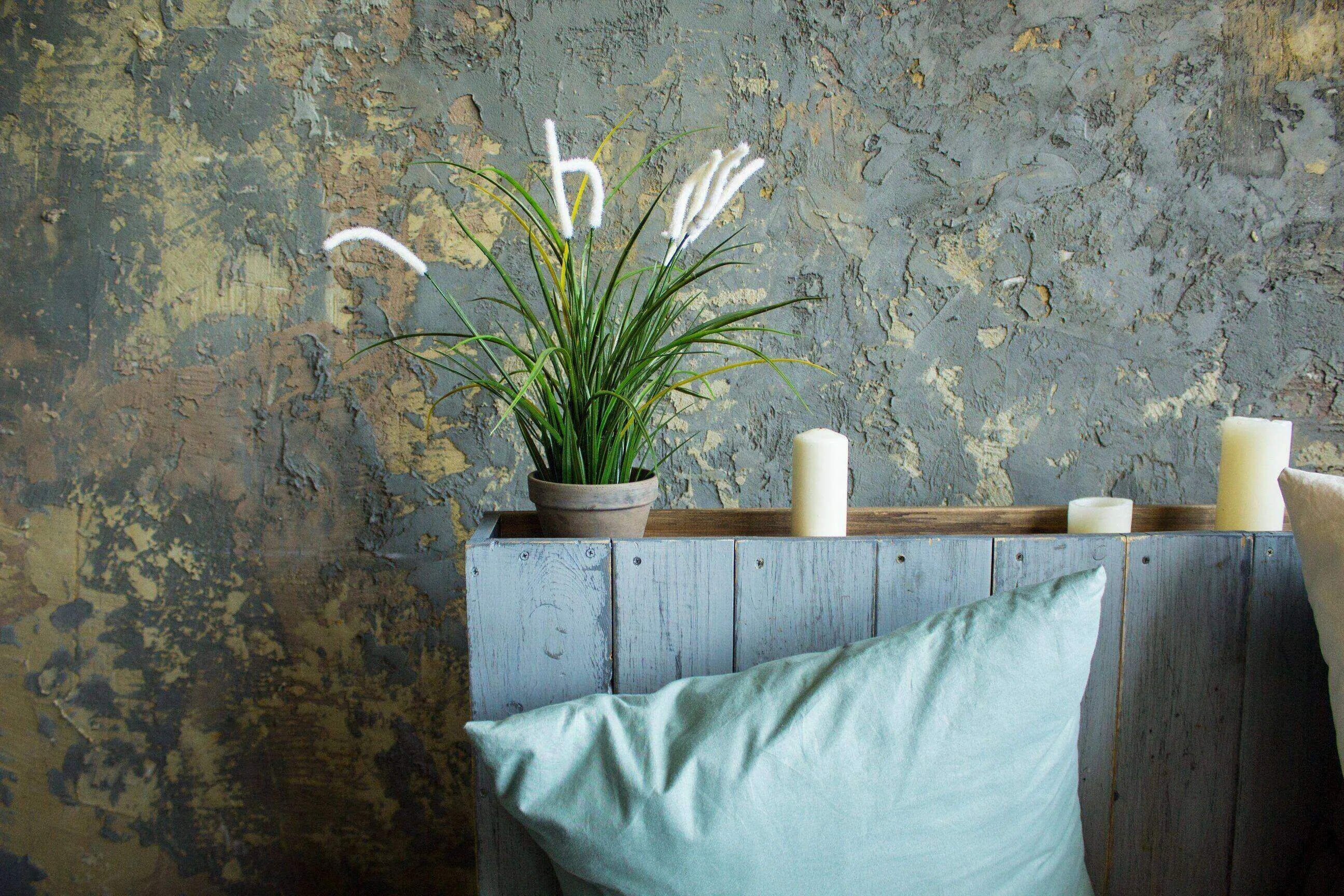 Textured wall with rustic finish, plant, candles, and light blue pillow on wooden shelf.