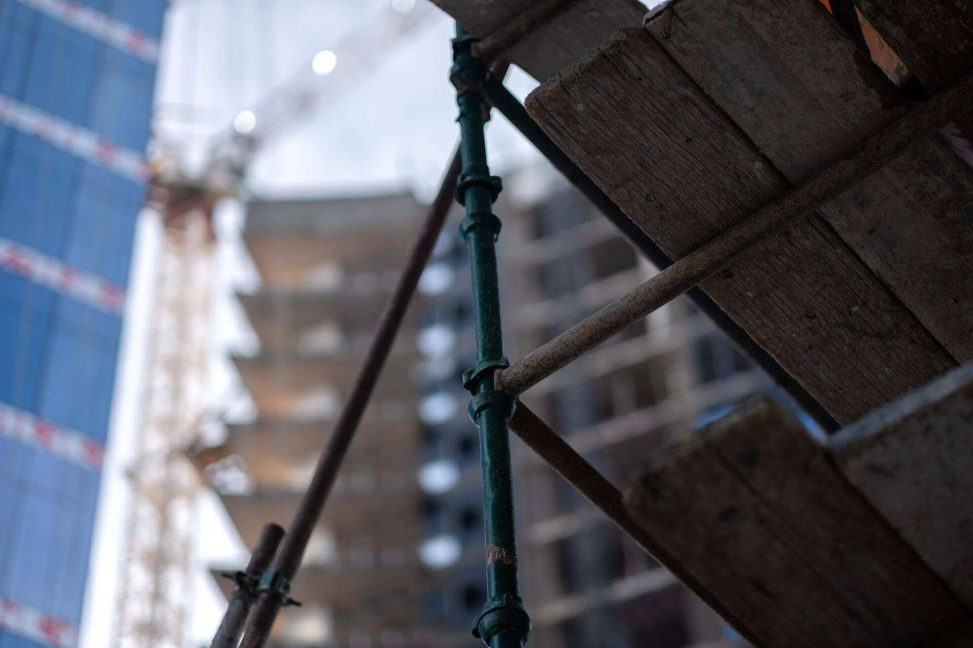 Close-up of scaffolding with wooden planks and metal poles at a construction site with a crane and high-rise building in the background.