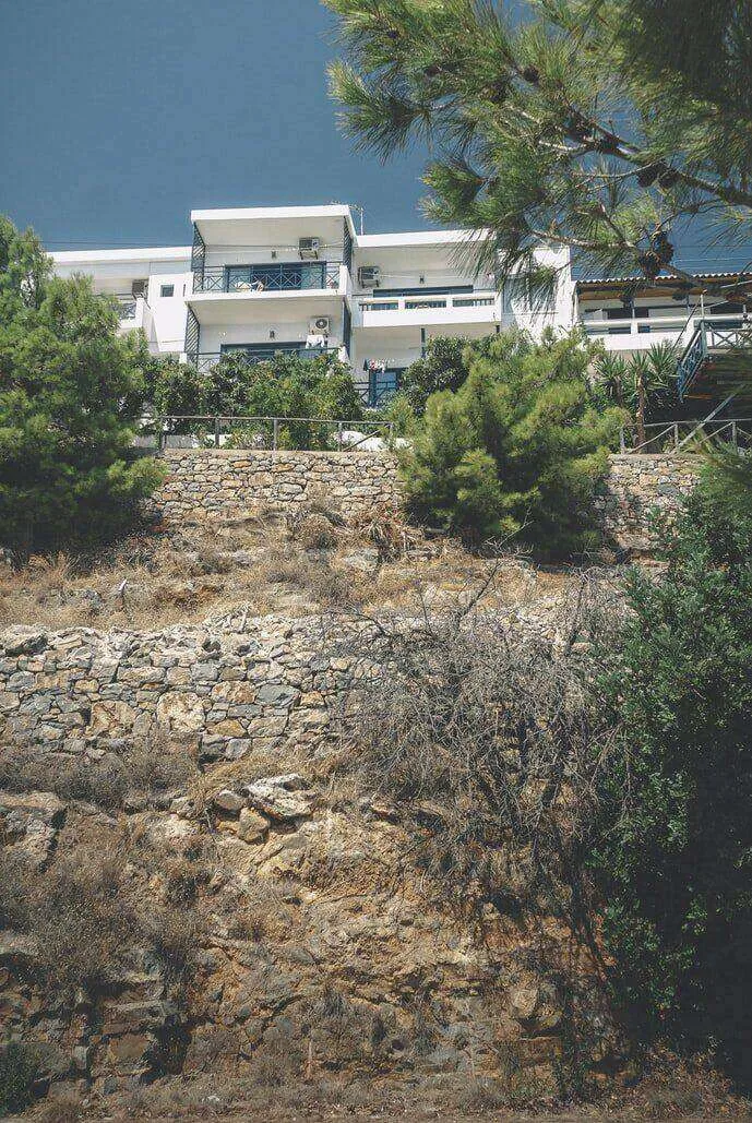 Stone retaining wall with a modern building above, surrounded by trees and shrubs.