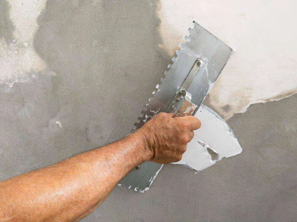 Close-up of a hand applying stucco to a wall with a notched trowel, showing repair work.