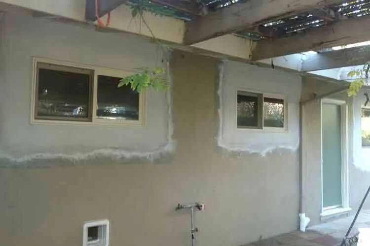 Residential exterior wall with two patched stucco areas around windows under a pergola.