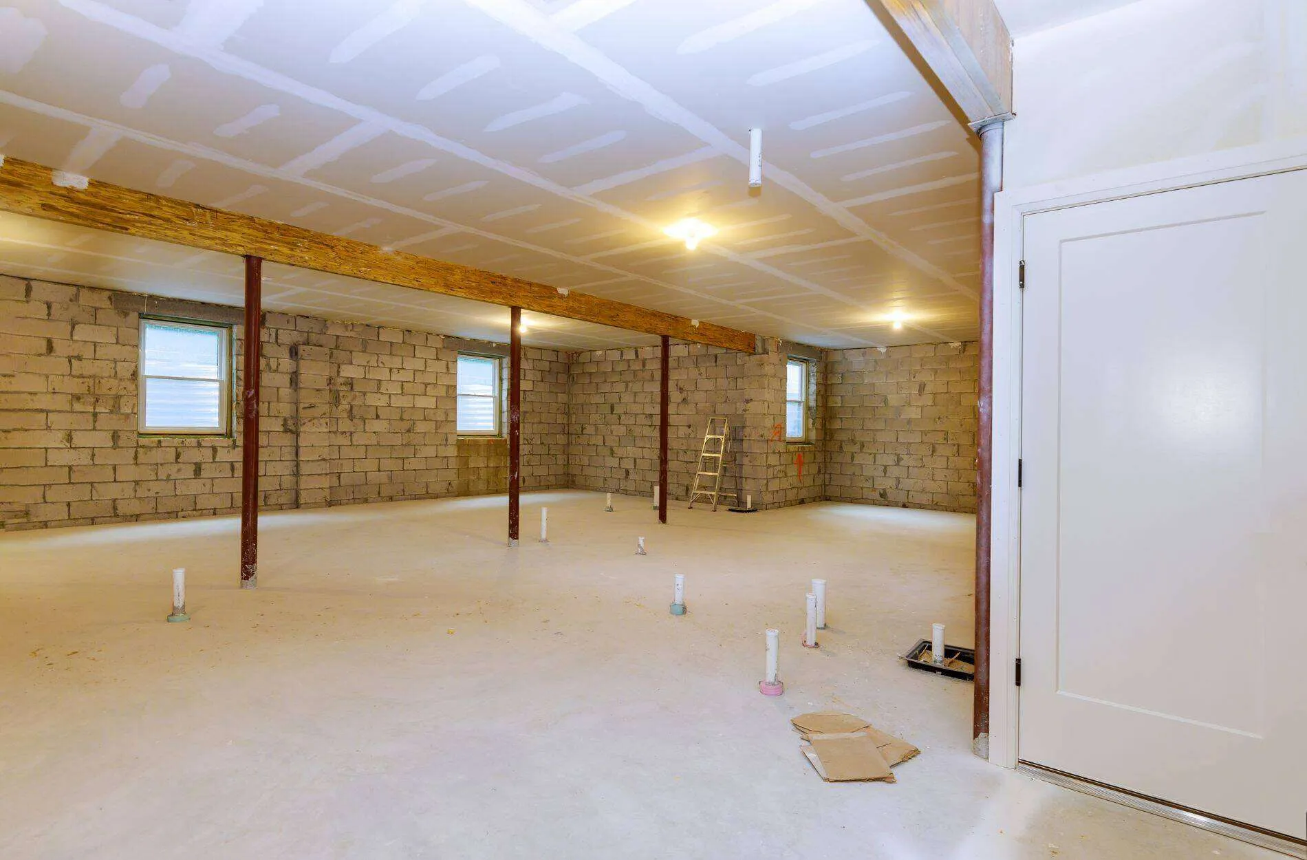 Unfinished basement with concrete block walls, exposed ceiling beams, and plumbing pipes protruding from the floor.