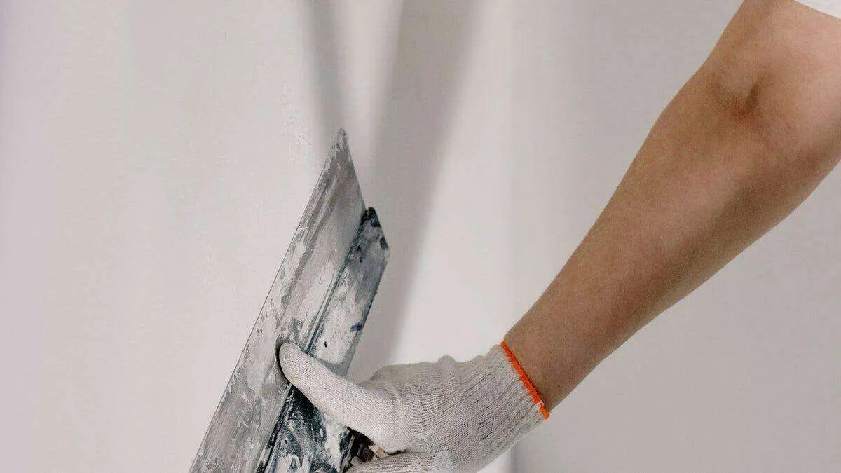 Person applying plaster to a wall with a trowel, wearing a glove.