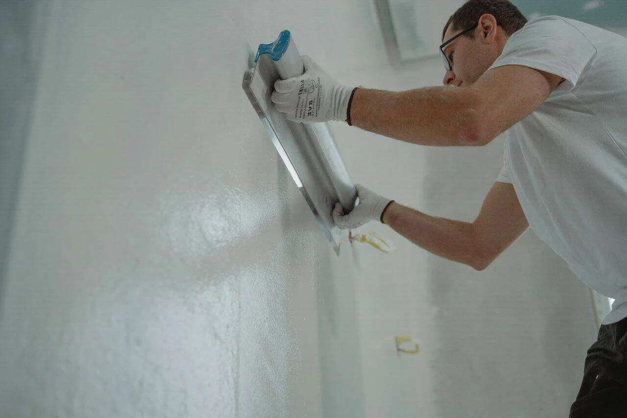 Person applying plaster to a wall with a trowel, wearing gloves and a white shirt.