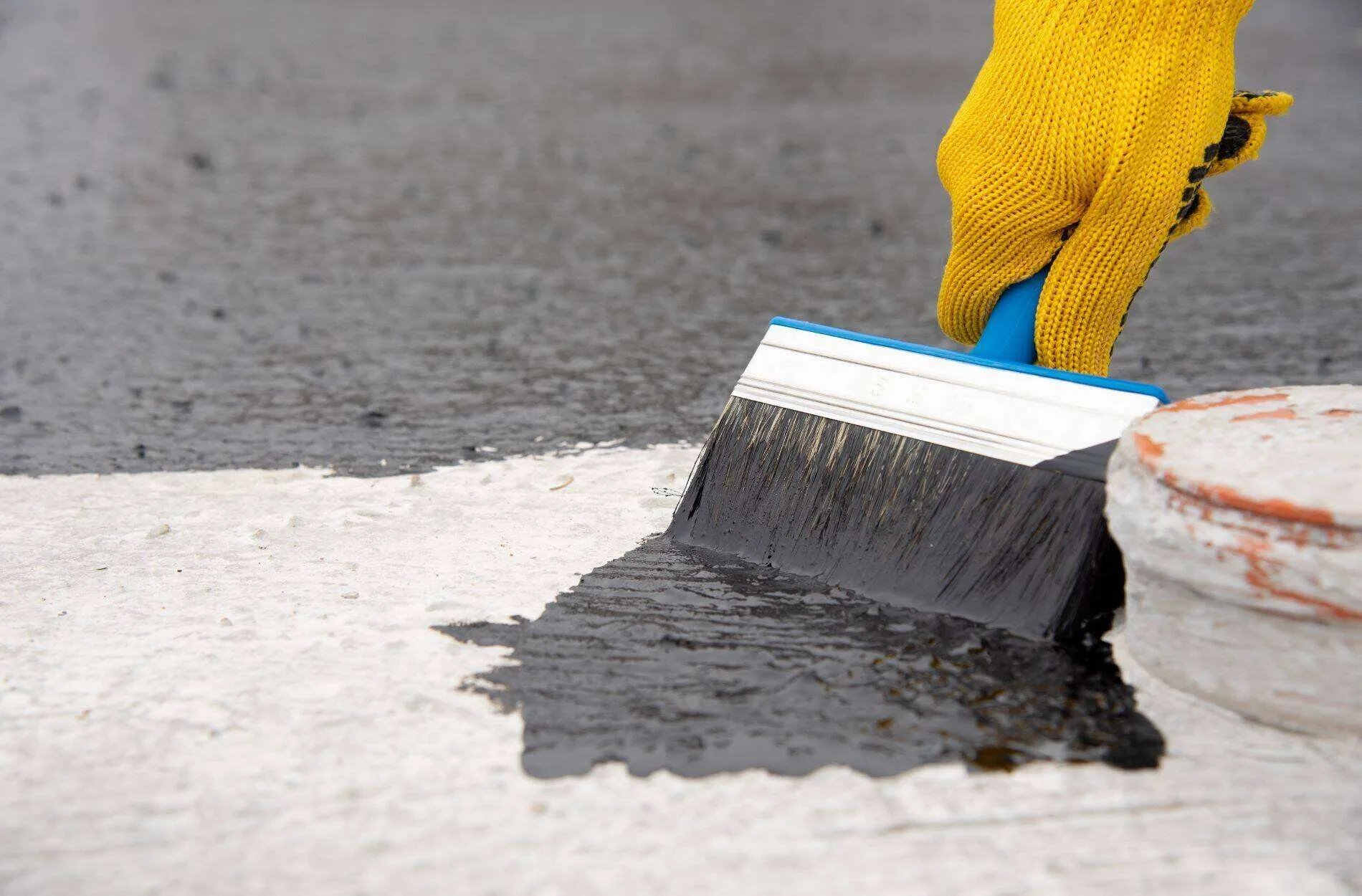 Person in yellow glove applying black waterproofing liquid to concrete with a brush.