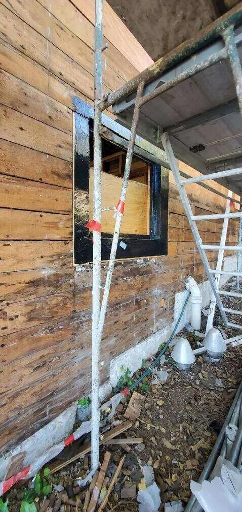 Scaffolding against a wooden wall with window opening and black waterproofing material.