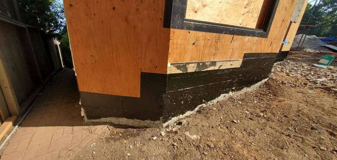 Building corner with plywood sheathing and black waterproofing membrane at the base, surrounded by bare soil.