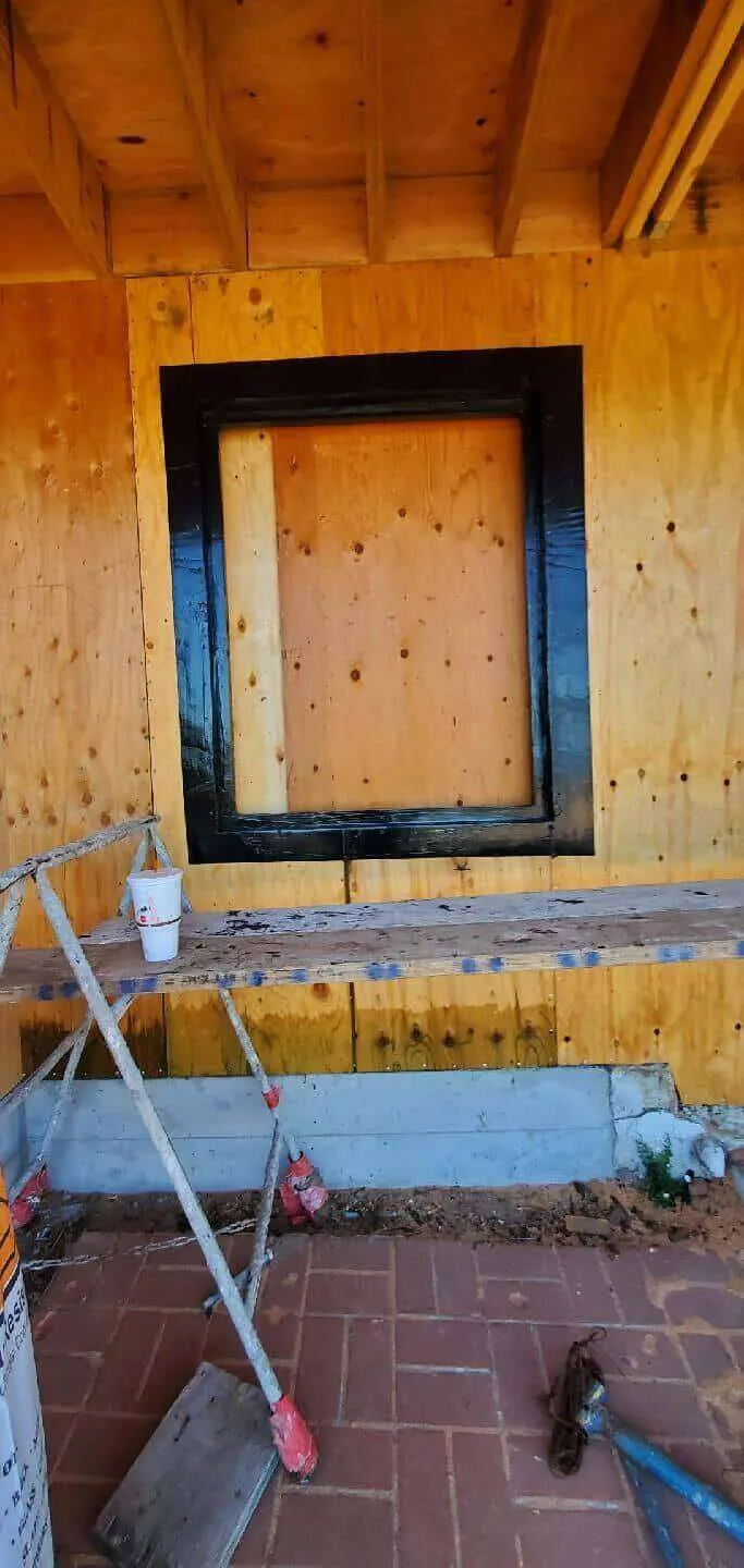Construction site with plywood walls and a window opening framed with black waterproofing material.