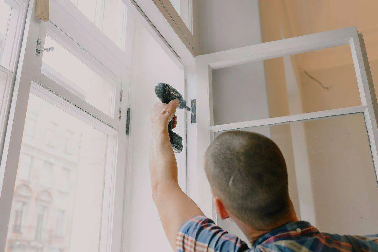 Person using a power drill on a window frame, indicating window installation or repair.