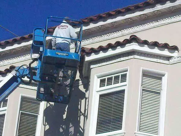 Worker on lift patching exterior near window with decorative trim and tiled roof.