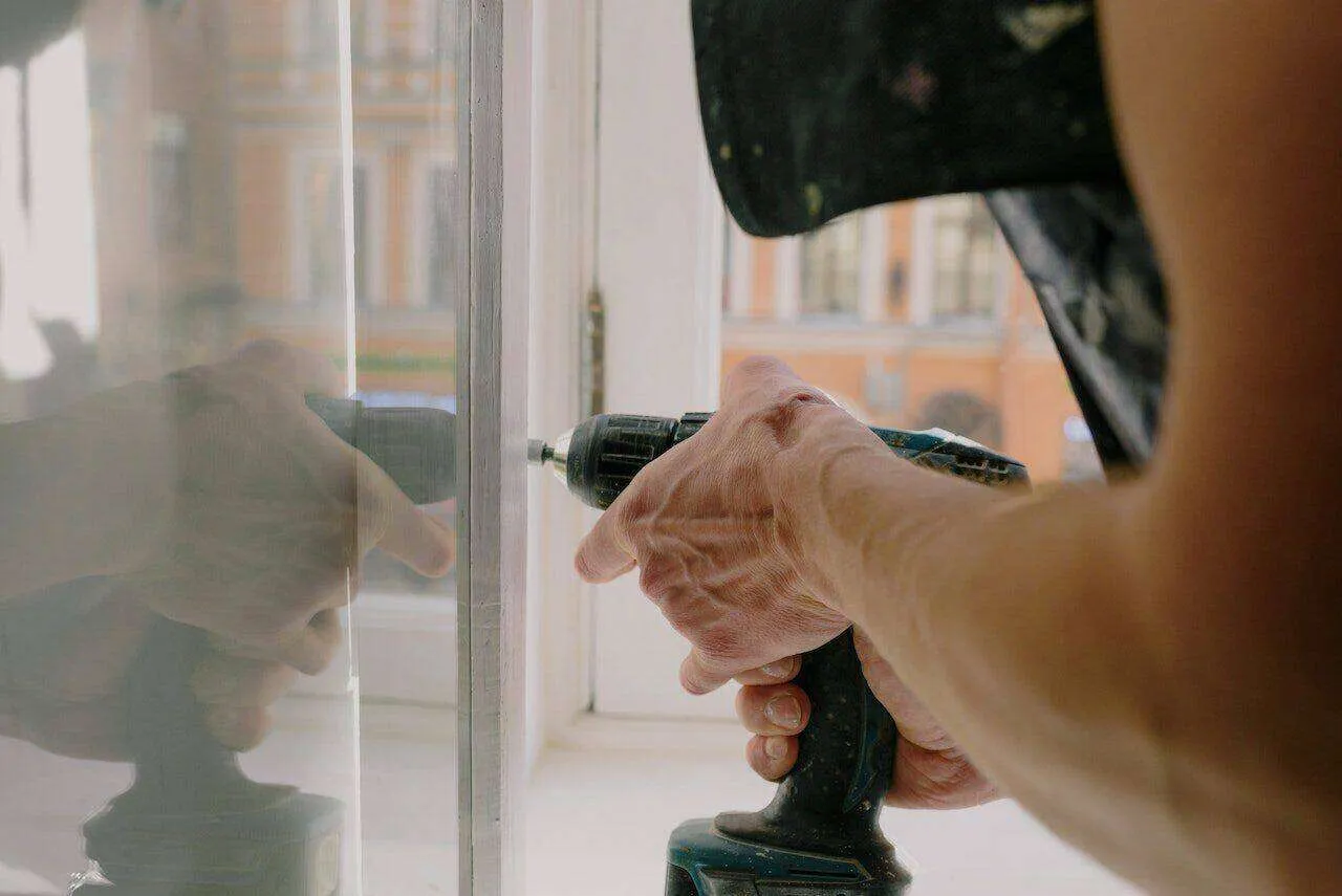 Person using a power drill on a window frame with reflection in glass.