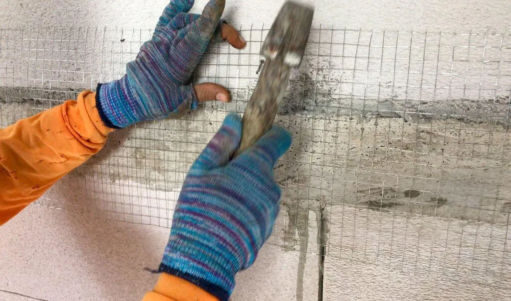 Person securing wire mesh to a wall with a hammer, wearing gloves.