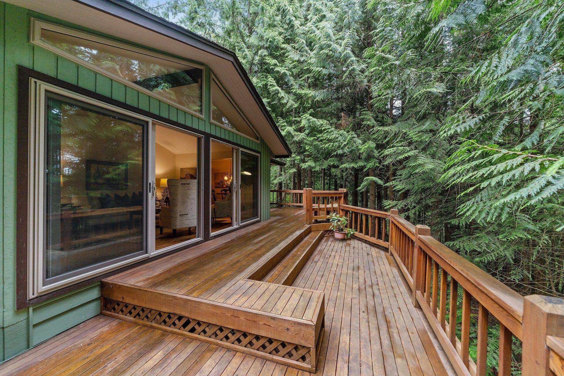 Wooden deck with railing, surrounded by trees, attached to a house with large glass doors.
