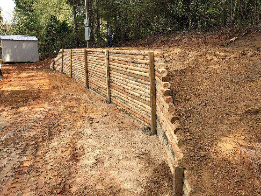 Wooden retaining wall in a yard with soil and a cleared path, surrounded by trees.