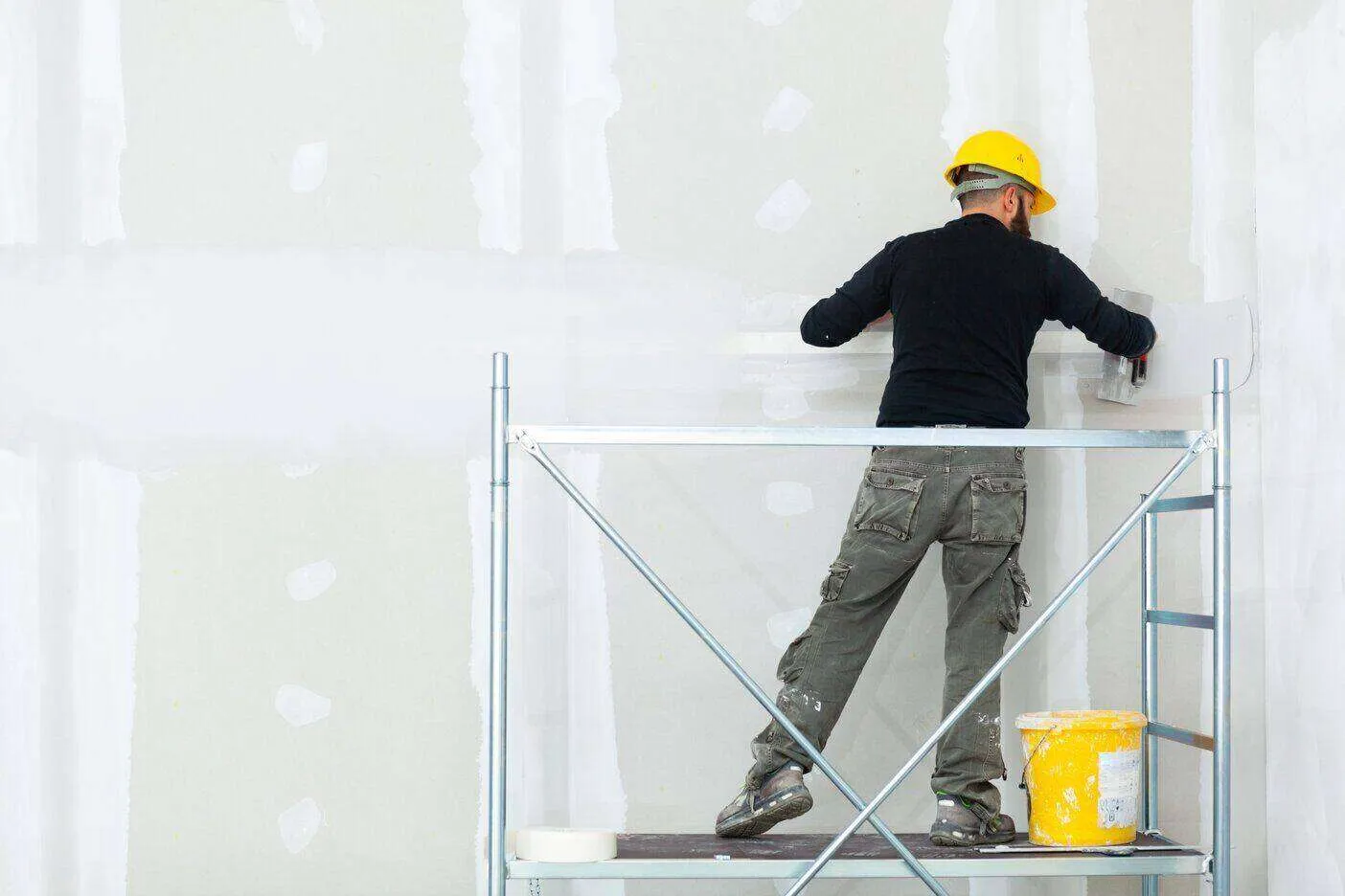 Worker in yellow hard hat plastering a gypsum board wall on scaffolding with a plaster bucket nearby.